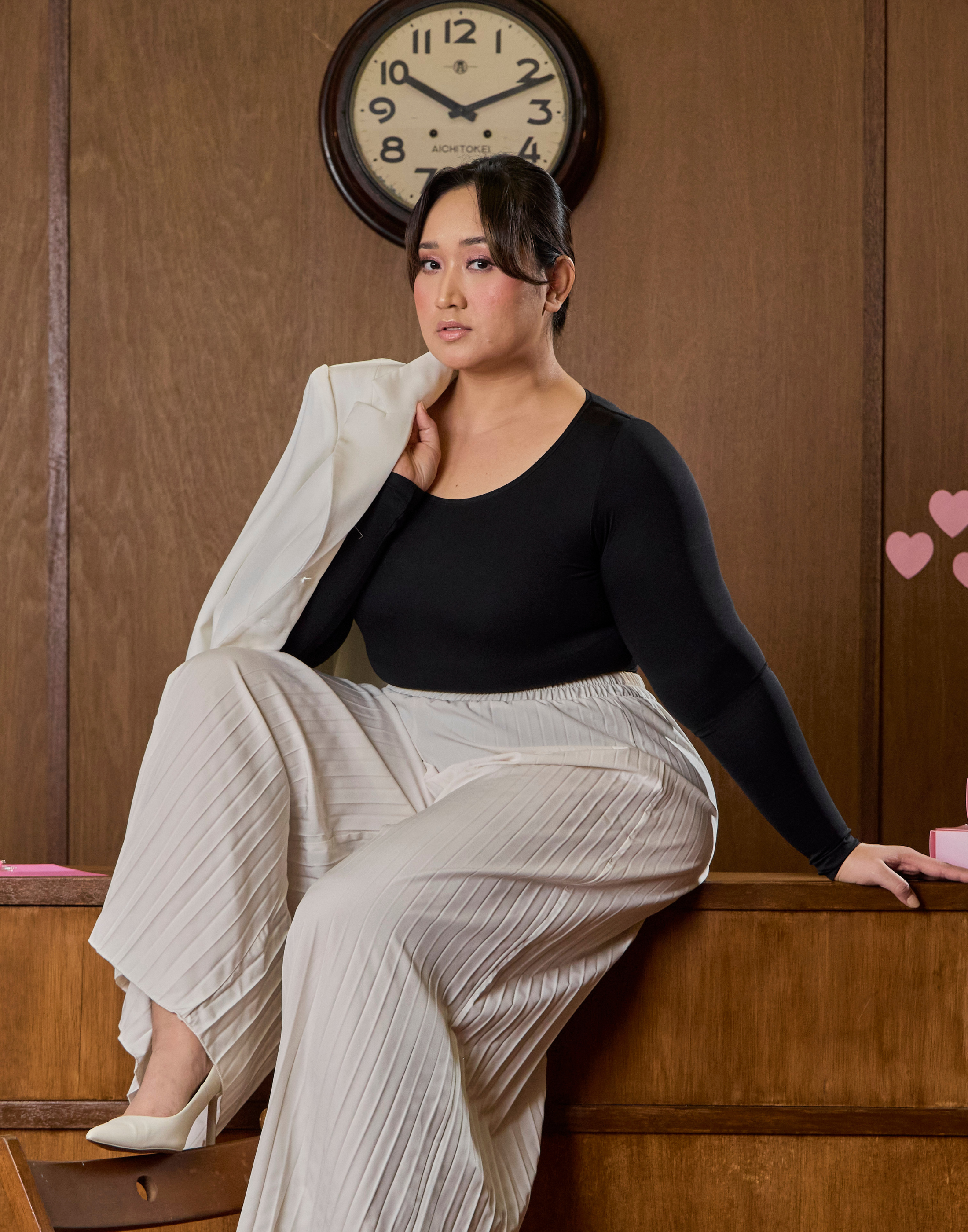 A person wearing a black arms shaping bodysuit with long sleeves, paired with light-colored striped trousers. The individual is seated, posing with one hand extended, in a setting with wooden paneling and a decorative clock on the wall.